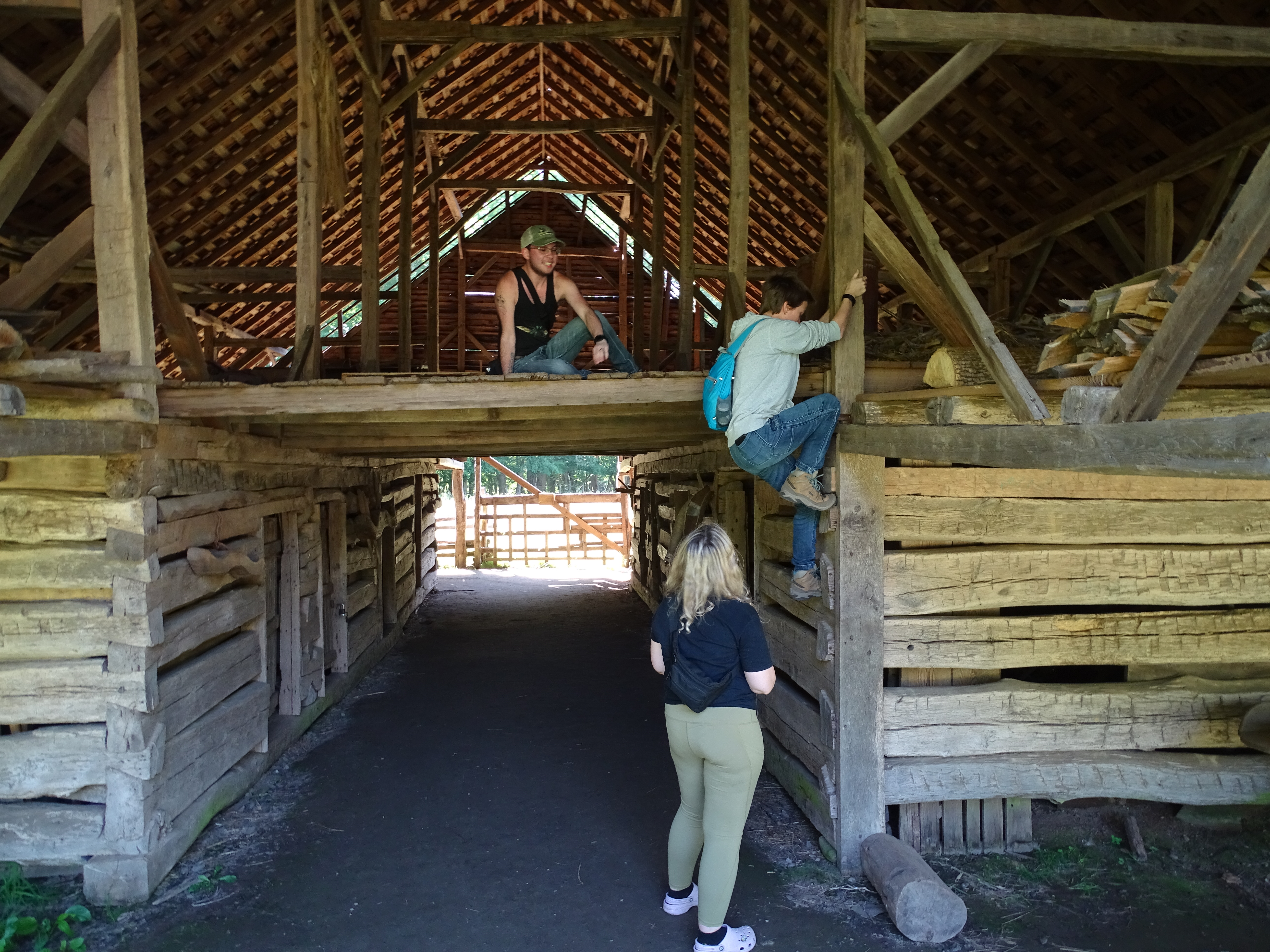 looking at a barn with a hay loft. One person is climbing up a wall to get to the loft, one person is sitting in the loft, and one person is standing on the ground watching the climber.
